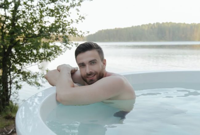 Shirtless man enjoying relaxation in hot tub by scenic lake, smiling and embracing leisure outdoors.