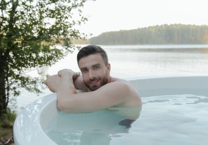 Shirtless man enjoying relaxation in hot tub by scenic lake, smiling and embracing leisure outdoors.